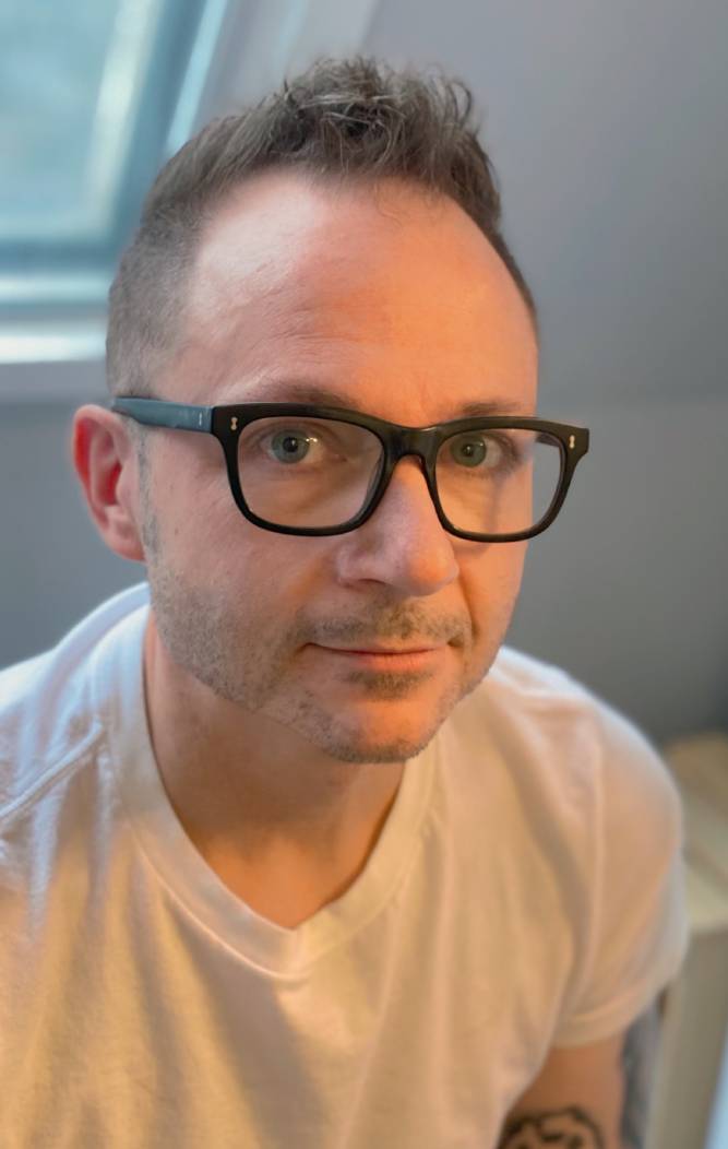 A photograph of set designer Douglas Tiller in a white t-shirt and dark framed glasses.