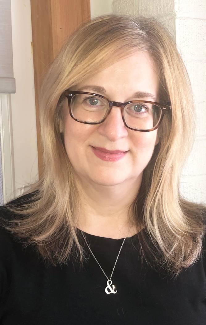 A photograph of producer and moss theatre collective member, Lori Delorme. Wearing a black top, dark framed glasses, and an ampersand necklace.